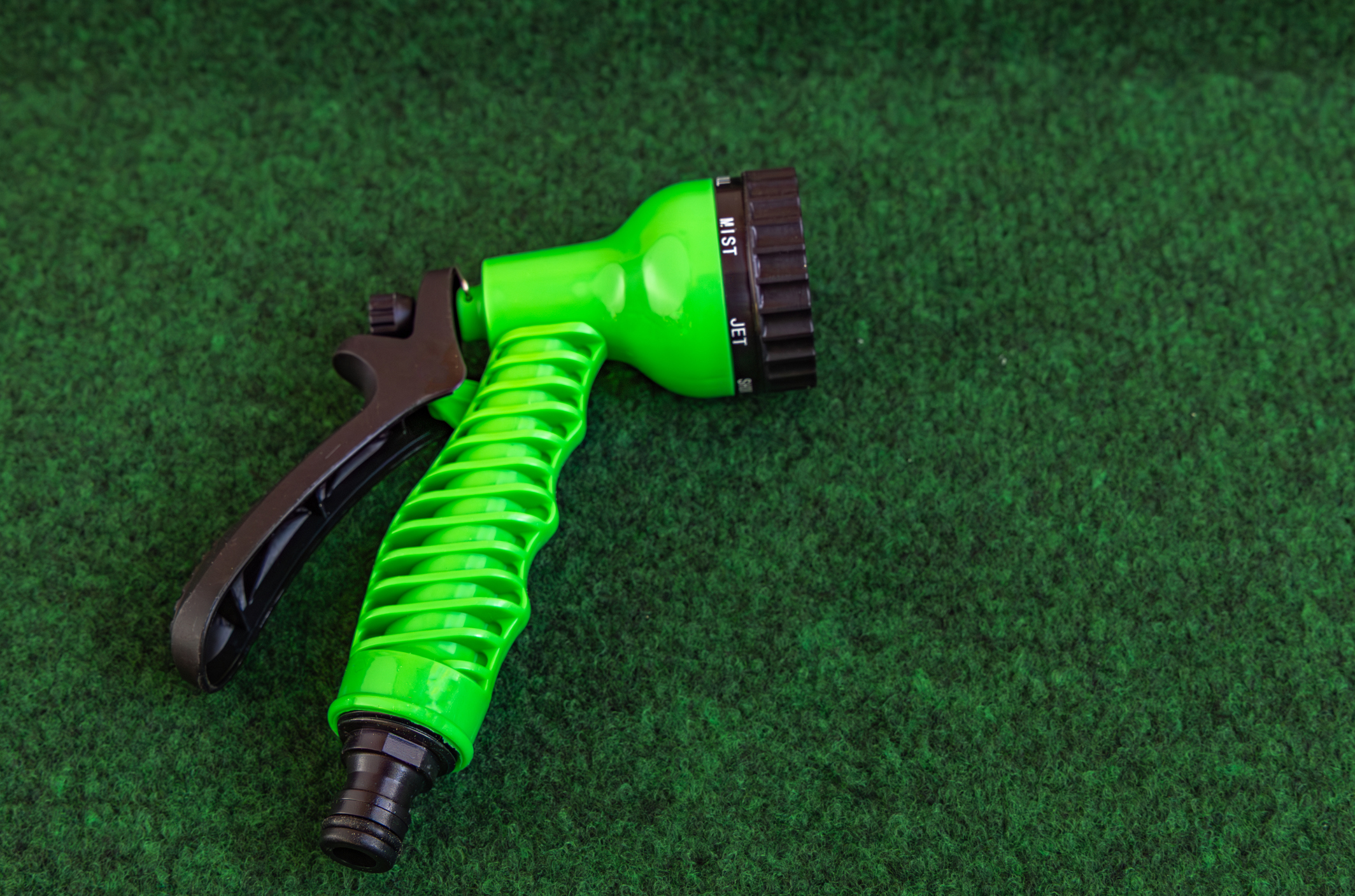 The green nozzle of a hose rests on artificial grass for how-to-clean-and-maintain-artificial-turf blog.